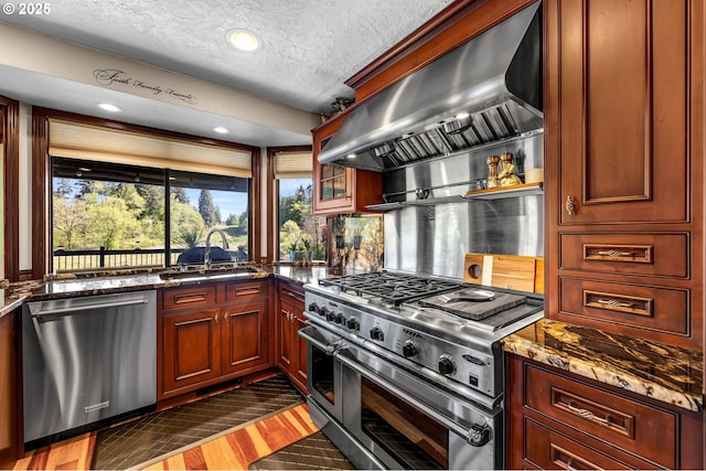kitchen with stainless steel appliances, a sink, wall chimney exhaust hood, dark stone counters, and a textured ceiling