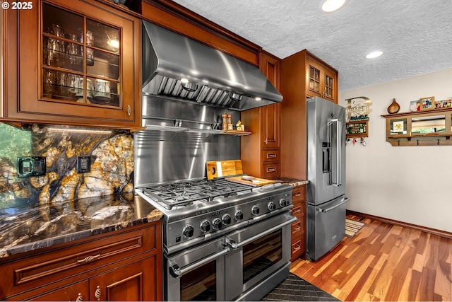 kitchen with high end appliances, wall chimney range hood, decorative backsplash, wood finished floors, and glass insert cabinets