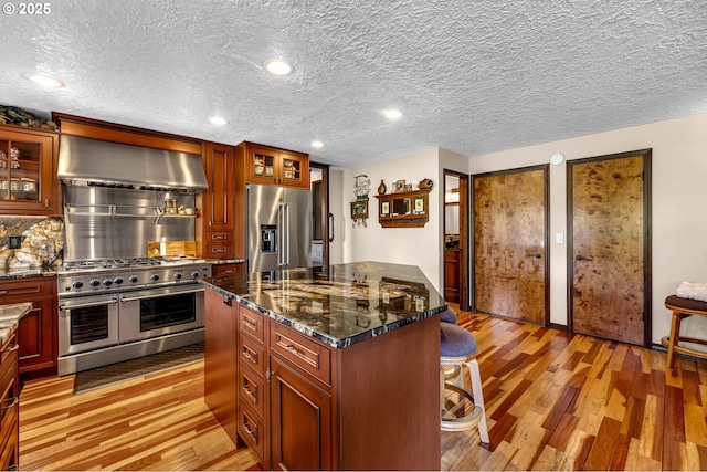 kitchen with high quality appliances, wall chimney exhaust hood, light wood-type flooring, a kitchen breakfast bar, and decorative backsplash