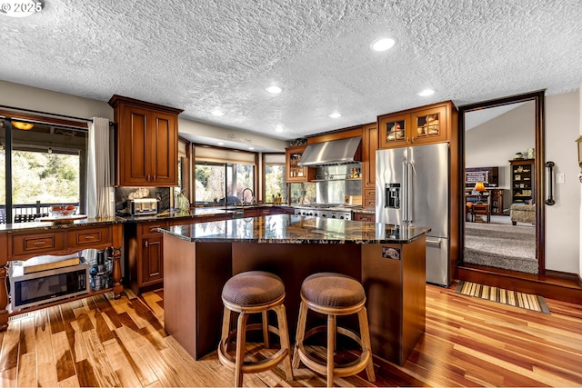 kitchen featuring appliances with stainless steel finishes, wall chimney exhaust hood, a textured ceiling, light wood finished floors, and glass insert cabinets