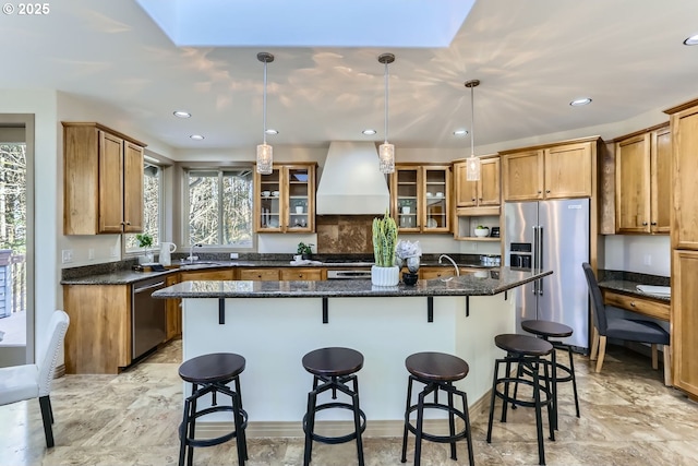 kitchen with premium range hood, appliances with stainless steel finishes, dark stone countertops, hanging light fixtures, and a kitchen island with sink