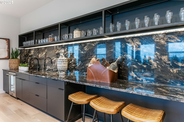 bar with backsplash, dark stone countertops, light hardwood / wood-style flooring, and sink