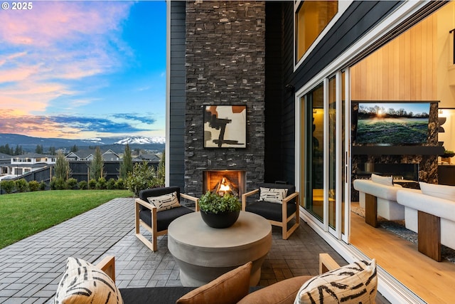 patio terrace at dusk with a mountain view, an outdoor stone fireplace, and a yard