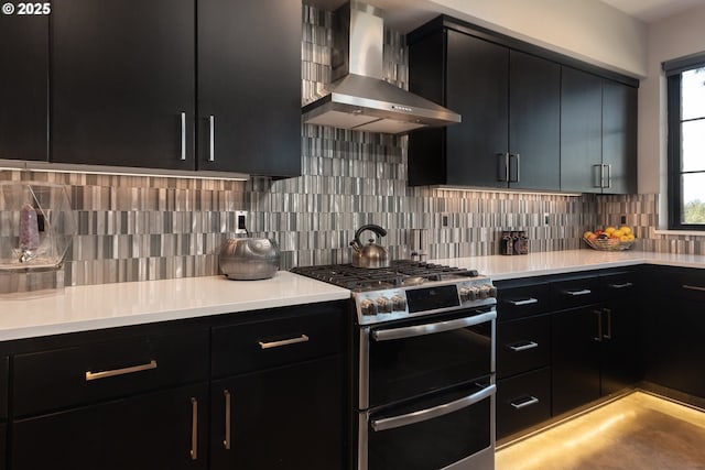kitchen featuring double oven range, wall chimney range hood, dark cabinetry, light countertops, and decorative backsplash