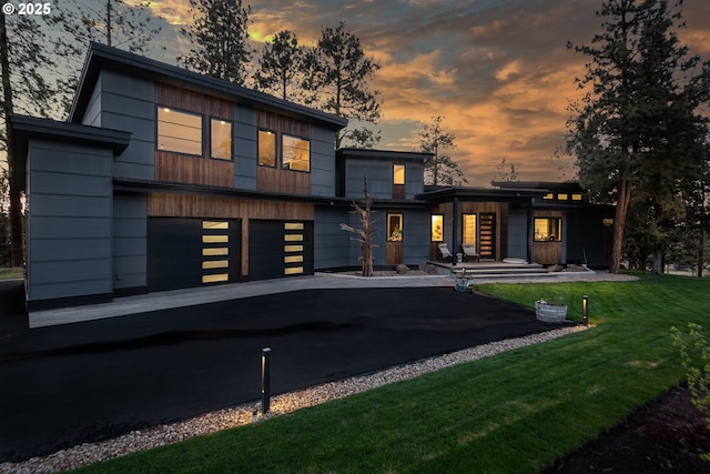 back of house at dusk featuring an attached garage, asphalt driveway, and a lawn