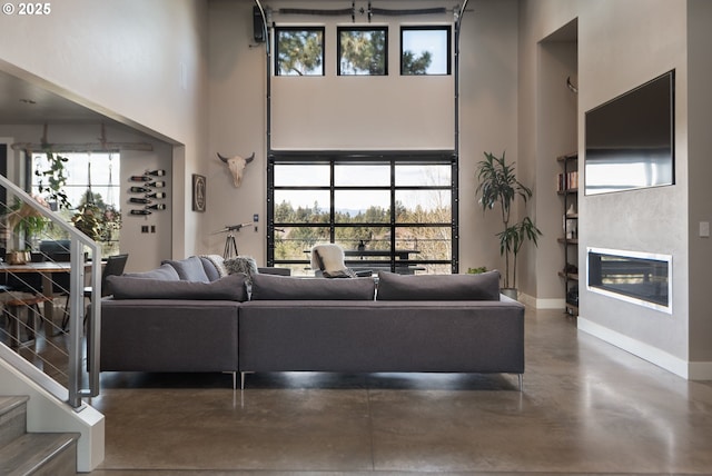 living area with a towering ceiling, concrete flooring, stairs, baseboards, and a glass covered fireplace