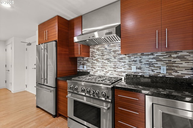 kitchen with stainless steel appliances, tasteful backsplash, brown cabinetry, wall chimney range hood, and dark stone counters