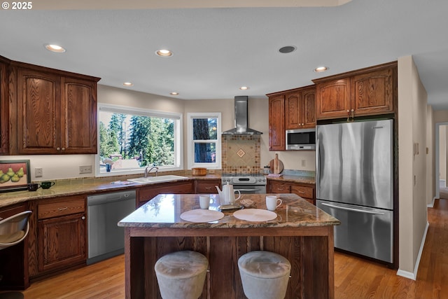 kitchen featuring appliances with stainless steel finishes, wall chimney range hood, light wood-style flooring, dark stone countertops, and recessed lighting