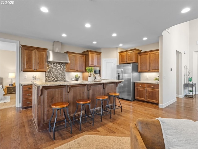 kitchen with wall chimney exhaust hood, stainless steel fridge, a kitchen breakfast bar, hardwood / wood-style flooring, and a kitchen island with sink