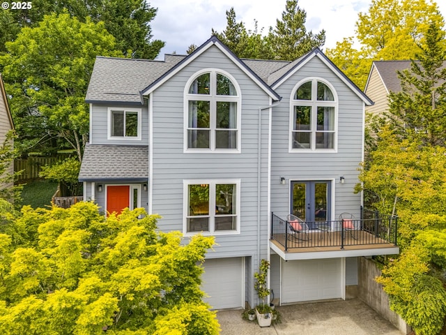 rear view of property featuring a shingled roof, french doors, a garage, driveway, and a balcony