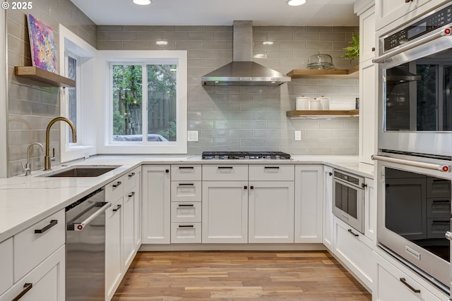 kitchen with open shelves, wall chimney range hood, appliances with stainless steel finishes, a sink, and light wood-style flooring