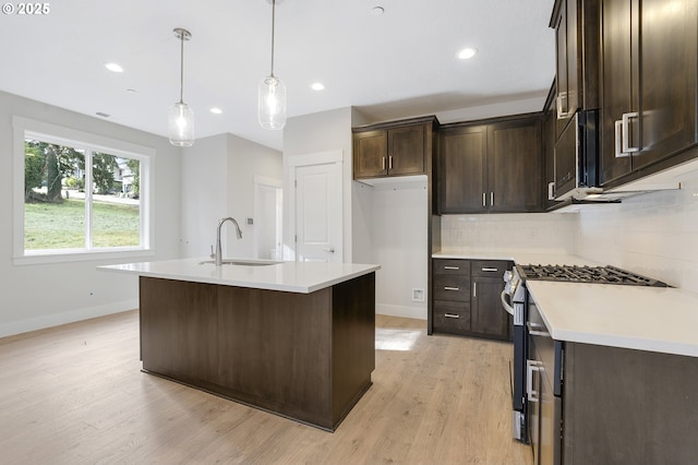 kitchen featuring stainless steel range with gas cooktop, an island with sink, sink, backsplash, and hanging light fixtures