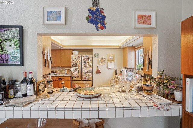 kitchen with freestanding refrigerator, tile counters, a peninsula, and a tray ceiling
