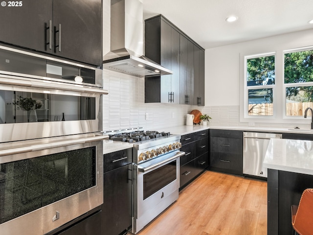 kitchen featuring wall chimney exhaust hood, sink, light hardwood / wood-style flooring, appliances with stainless steel finishes, and decorative backsplash