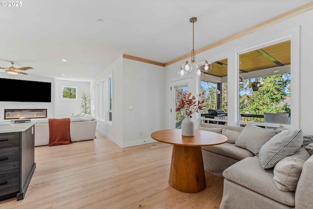 living room with light wood finished floors, crown molding, baseboards, ceiling fan with notable chandelier, and a glass covered fireplace