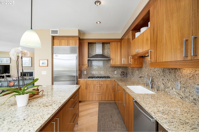 kitchen with wall chimney exhaust hood, stainless steel appliances, a sink, brown cabinetry, and light stone counters