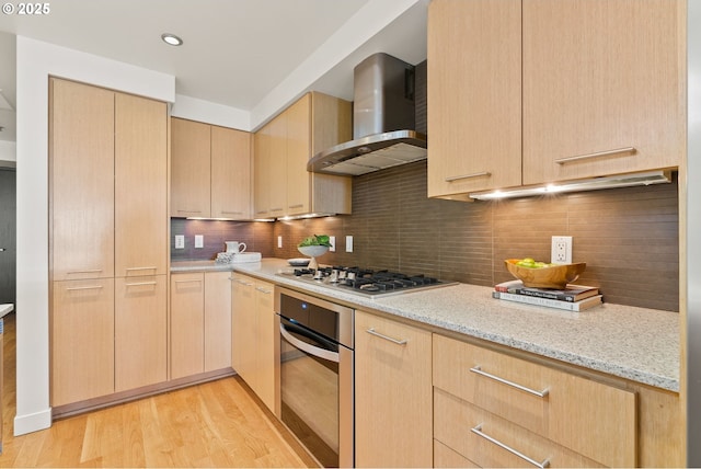 kitchen with light brown cabinetry, light stone counters, light wood-style floors, wall chimney exhaust hood, and stainless steel appliances