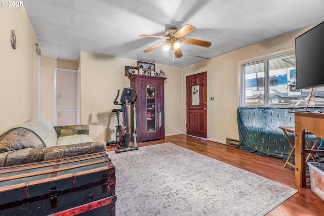 living area with a baseboard heating unit, ceiling fan, a textured ceiling, wood finished floors, and baseboards