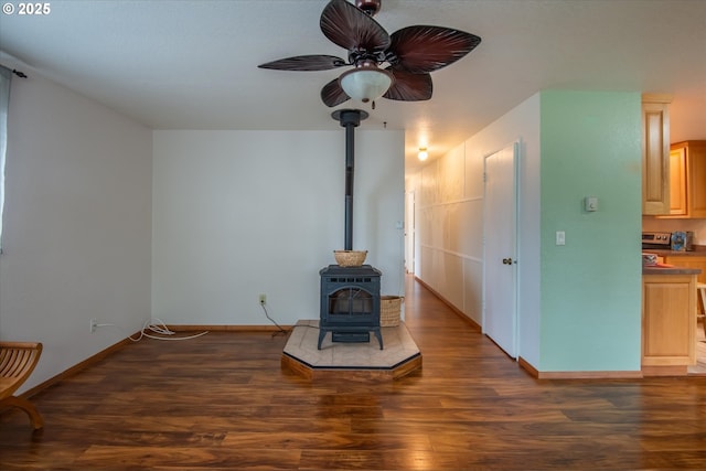 living room featuring a wood stove, dark wood-style floors, baseboards, and a ceiling fan