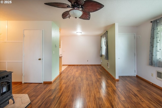 unfurnished living room featuring dark wood-type flooring, a wood stove, ceiling fan, and baseboards