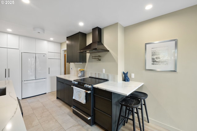 kitchen featuring wall chimney exhaust hood, a kitchen bar, electric range, built in fridge, and white cabinets