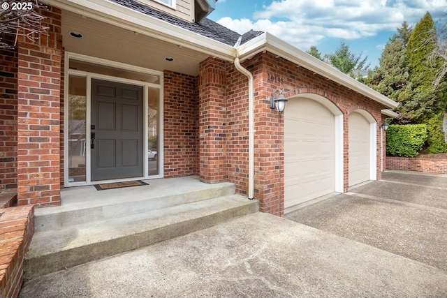 view of exterior entry with a garage, brick siding, driveway, and roof with shingles