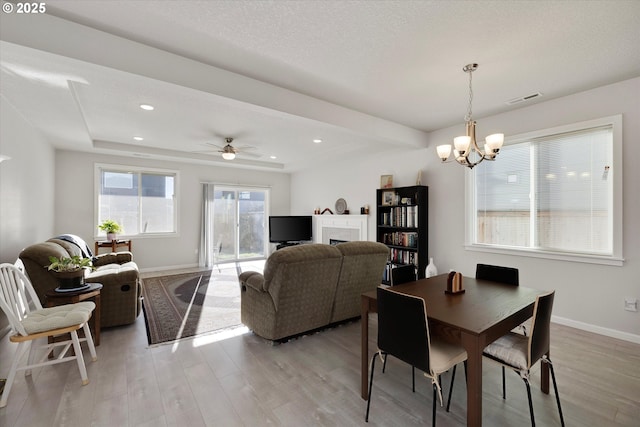 dining space with ceiling fan with notable chandelier, light hardwood / wood-style floors, a raised ceiling, and a tiled fireplace