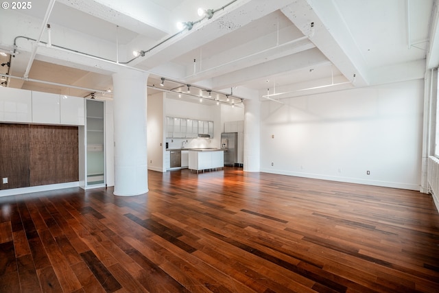 unfurnished living room featuring beamed ceiling, rail lighting, dark wood-type flooring, and baseboards