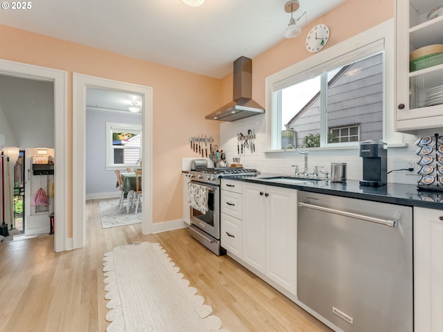 kitchen with stainless steel appliances, a sink, wall chimney range hood, dark countertops, and white cabinetry