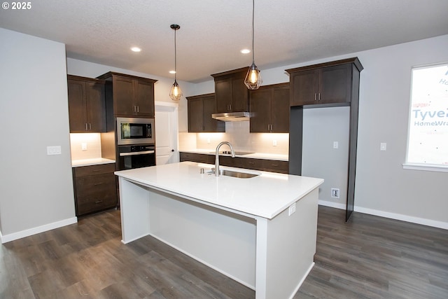 kitchen with sink, a center island with sink, built in microwave, decorative light fixtures, and stainless steel oven