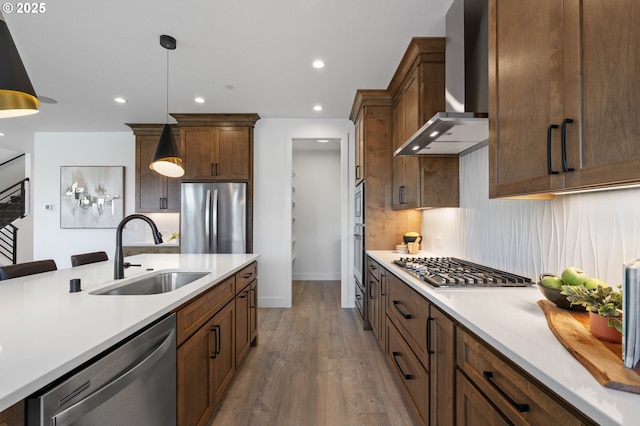 kitchen with stainless steel appliances, light countertops, hanging light fixtures, a sink, and wall chimney range hood