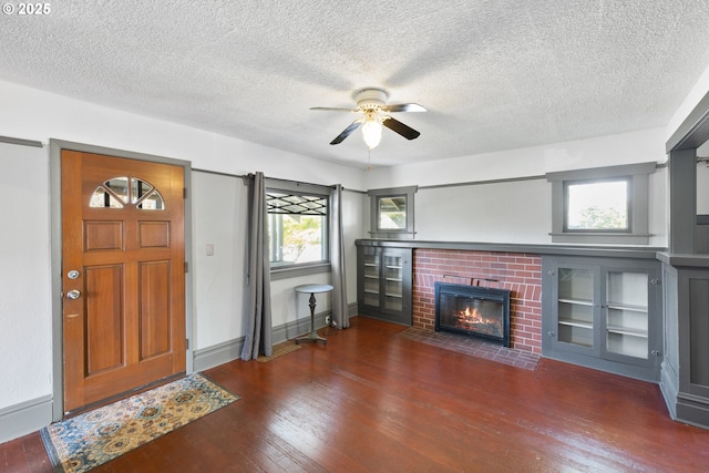 unfurnished living room with a textured ceiling, dark wood-style flooring, a ceiling fan, baseboards, and a brick fireplace