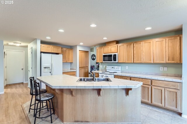 kitchen with tile countertops, light brown cabinetry, a sink, white appliances, and a kitchen breakfast bar