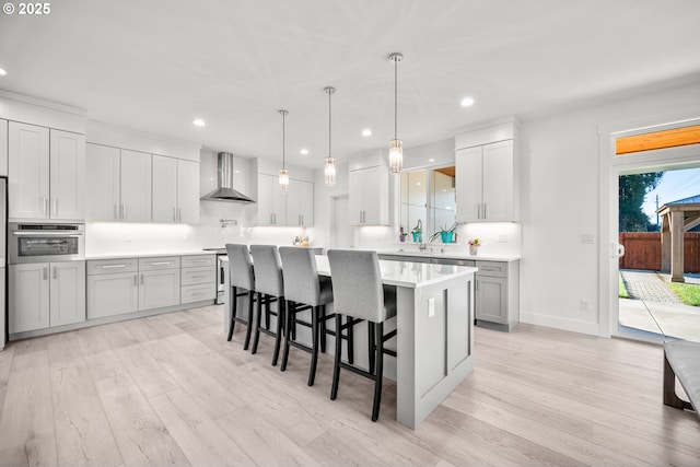 kitchen with wall chimney range hood, a breakfast bar, light countertops, stainless steel oven, and light wood-style flooring