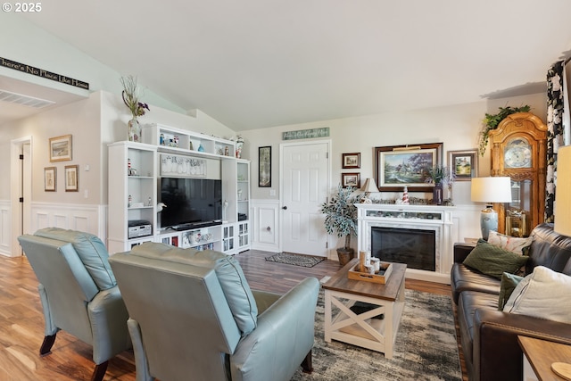 living area featuring wainscoting, wood finished floors, vaulted ceiling, a decorative wall, and a glass covered fireplace