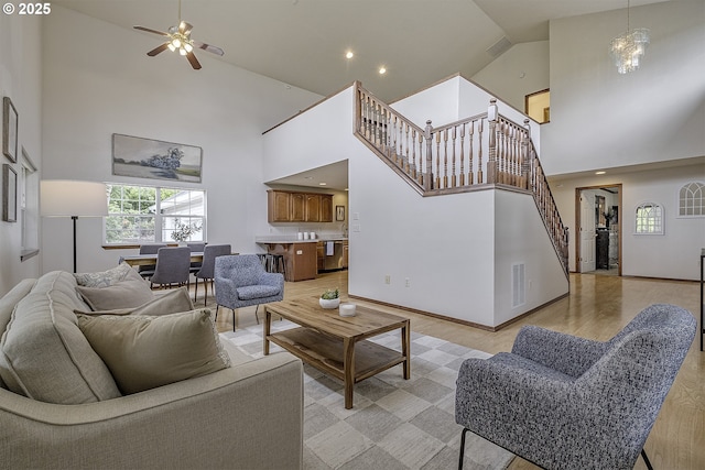 living room with baseboards, stairs, a high ceiling, light wood-style floors, and ceiling fan with notable chandelier