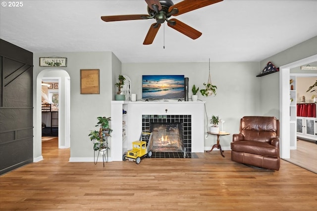 living room featuring a ceiling fan, baseboards, wood finished floors, arched walkways, and a tile fireplace