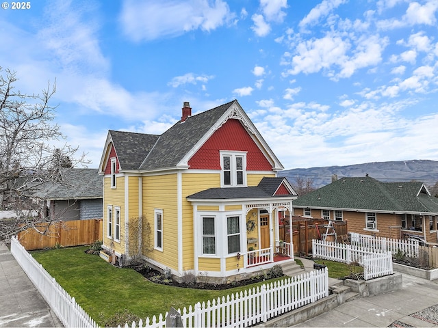 victorian house with a fenced front yard, a mountain view, a shingled roof, a front lawn, and a chimney