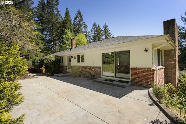 rear view of house with brick siding, roof with shingles, a chimney, and a patio