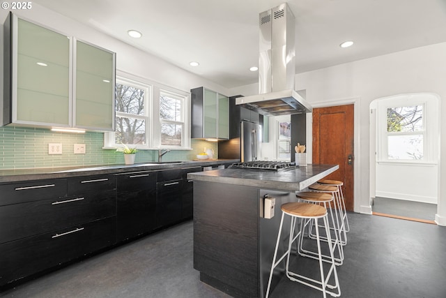 kitchen with island range hood, dark countertops, stainless steel gas stovetop, finished concrete floors, and a kitchen bar