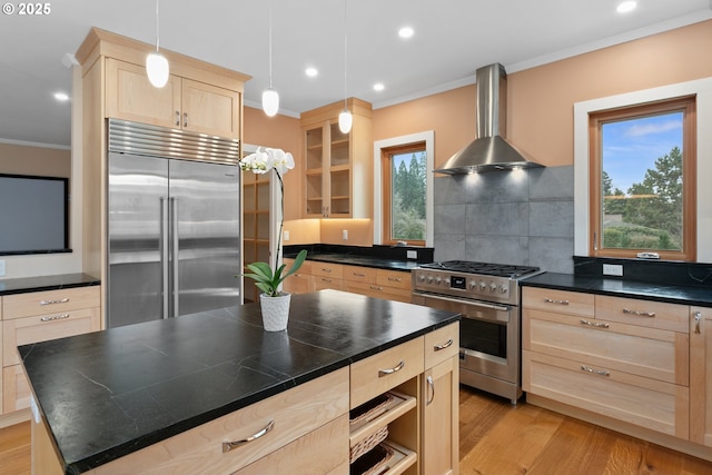 kitchen featuring dark countertops, wall chimney exhaust hood, a kitchen island, appliances with stainless steel finishes, and glass insert cabinets