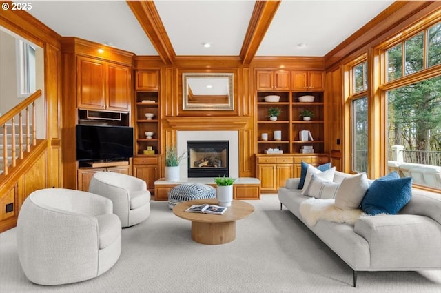 living room with built in shelves, a glass covered fireplace, light colored carpet, ornamental molding, and beamed ceiling