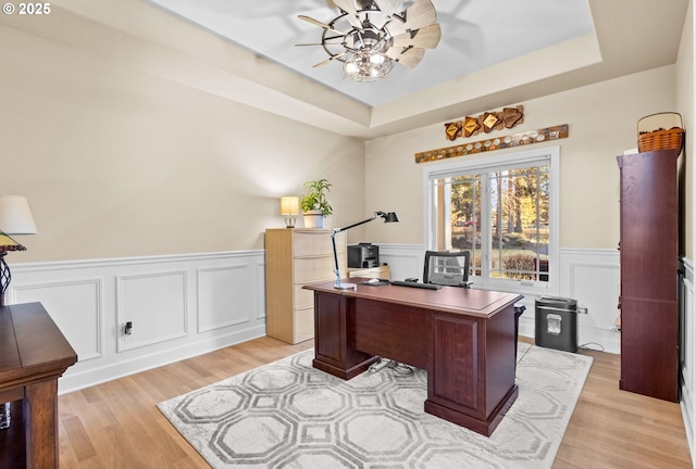 office space with a wainscoted wall, a raised ceiling, light wood-style flooring, and a ceiling fan