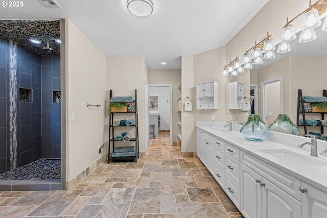 bathroom featuring stone tile floors, a sink, baseboards, double vanity, and walk in shower