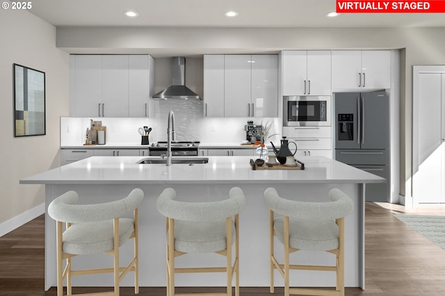 kitchen with white cabinets, wall chimney exhaust hood, stainless steel appliances, and a center island with sink