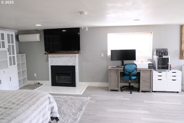 bedroom featuring an office area, a wall mounted AC, a glass covered fireplace, light wood finished floors, and baseboards