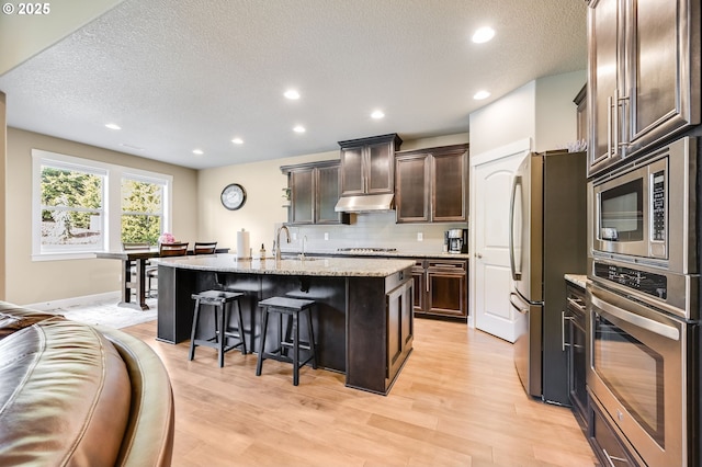 kitchen with appliances with stainless steel finishes, dark brown cabinets, light stone countertops, a breakfast bar, and a kitchen island with sink