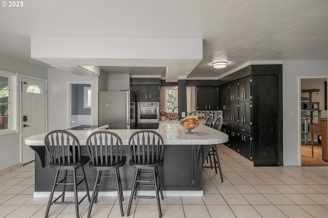 kitchen with appliances with stainless steel finishes, tile countertops, light tile patterned floors, a kitchen breakfast bar, and dark cabinetry