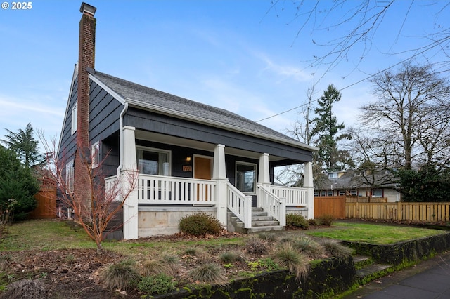 bungalow-style house with a chimney, a porch, roof with shingles, fence, and a front lawn