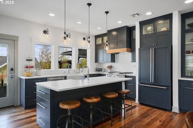 kitchen with high end refrigerator, dark wood finished floors, a kitchen island with sink, light countertops, and a sink